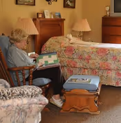 An older adult sitting in a chair reading near a bed with a floral quilt in a cozy, well-decorated bedroom.