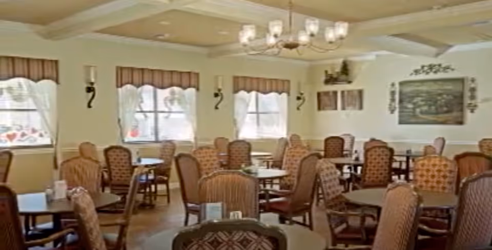 A dining room with multiple round tables surrounded by upholstered chairs. The room has large windows with curtains and valances, wall sconces, and a chandelier hanging from a coffered ceiling. There are framed pictures and decorative items on the walls.