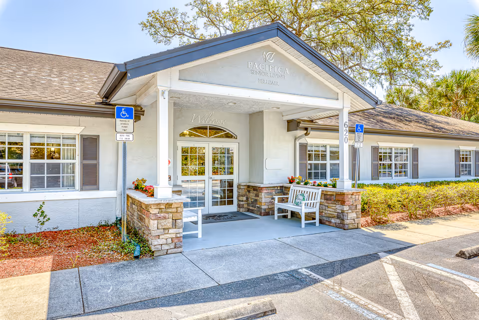 Exterior front entrance of a senior living facility named Pacifica Senior Living Belleair, featuring a covered porch with two white benches, stone pillars, and handicap parking signs on either side of the entrance.