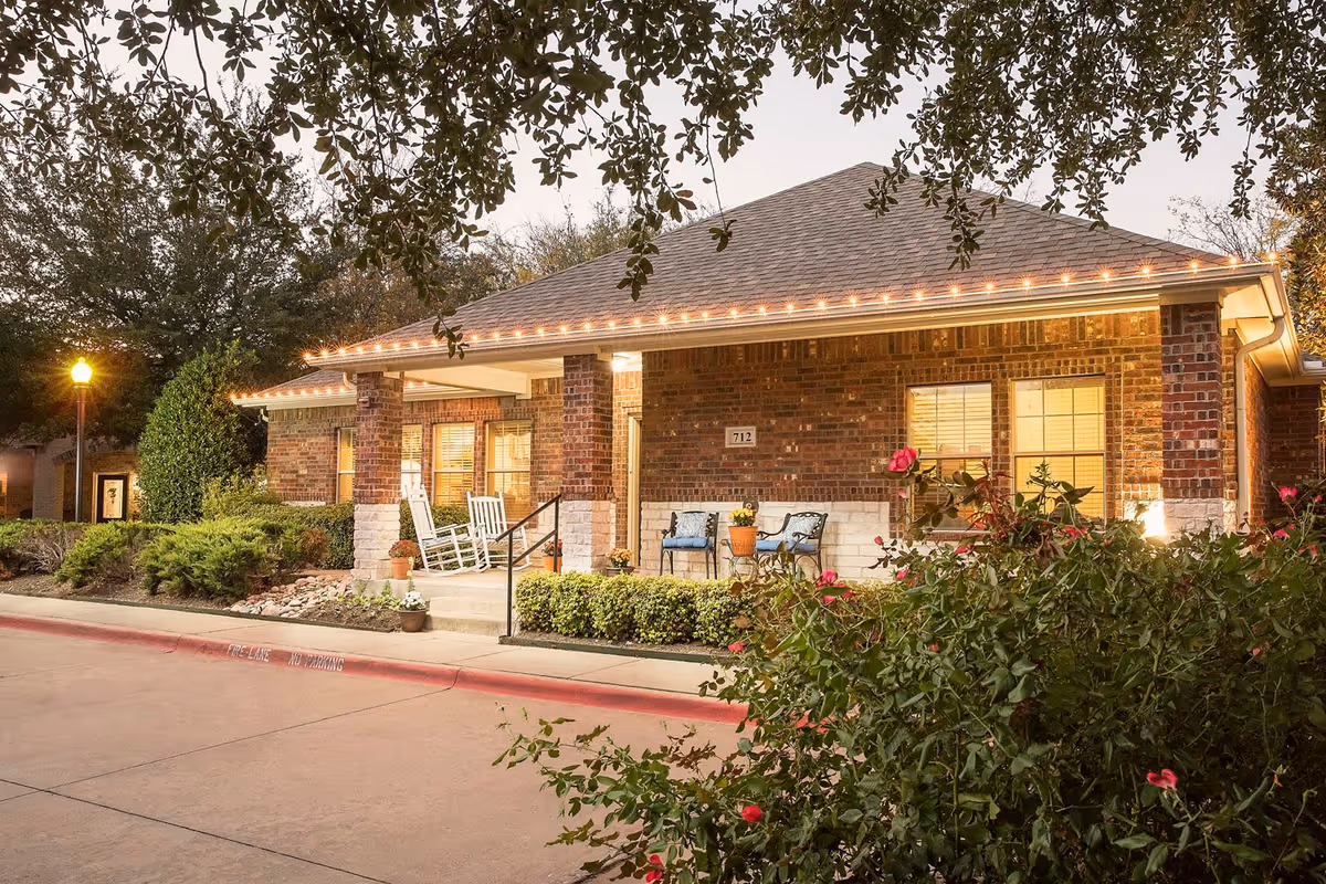 Exterior view of a single-story brick building with a covered porch featuring rocking chairs and a small table with chairs. The building is surrounded by well-maintained bushes and flowering plants, with a streetlamp visible on the left side and trees overhead.
