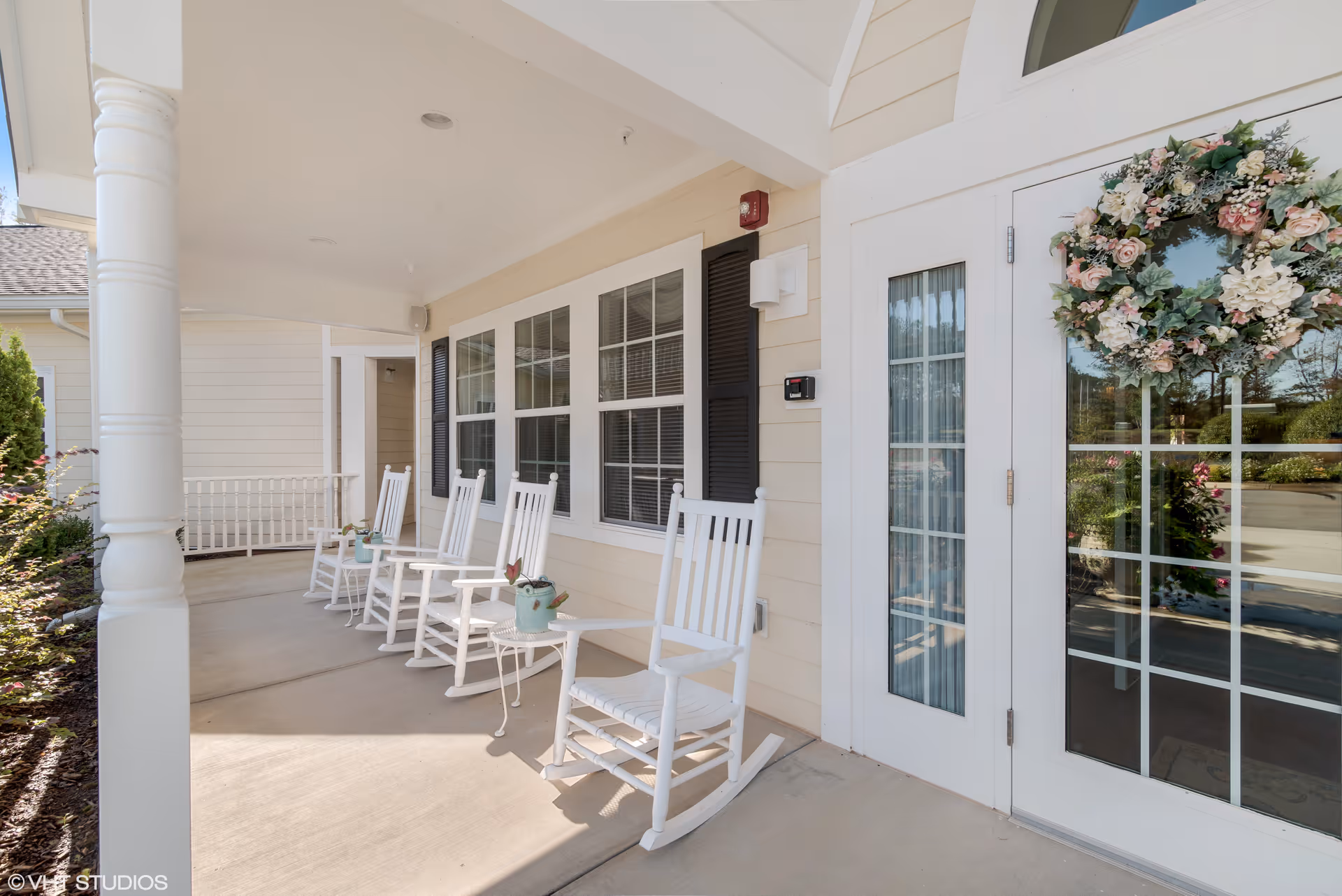 Covered porch area with white rocking chairs and small tables, decorated with potted plants. The porch is attached to a beige building with white trim and black shutters. A glass door with a floral wreath is visible on the right side.