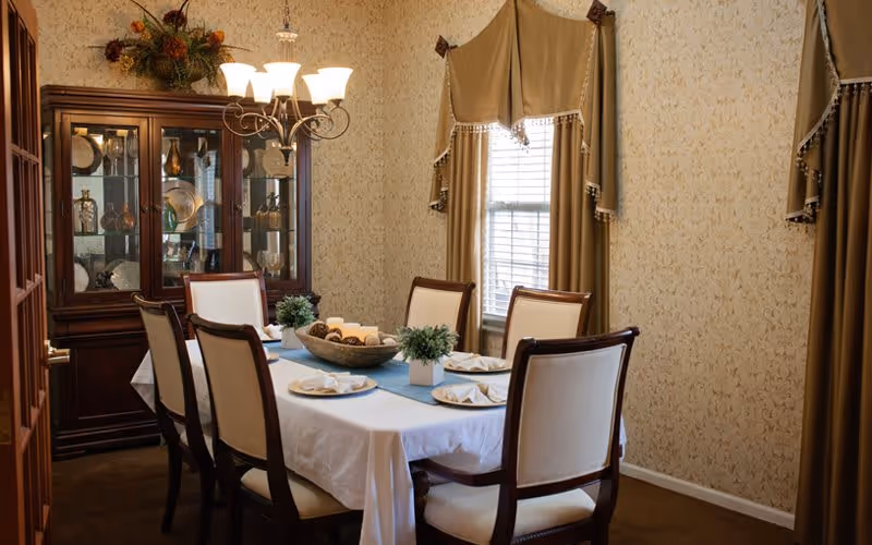 A formal dining room with a rectangular table covered with a white tablecloth and a blue runner. Six upholstered chairs surround the table, which is set with plates and folded napkins. A wooden china cabinet with glass doors displays decorative dishes and glassware. The room has beige patterned wallpaper, two windows with gold curtains, and a chandelier with five lights hanging above the table.