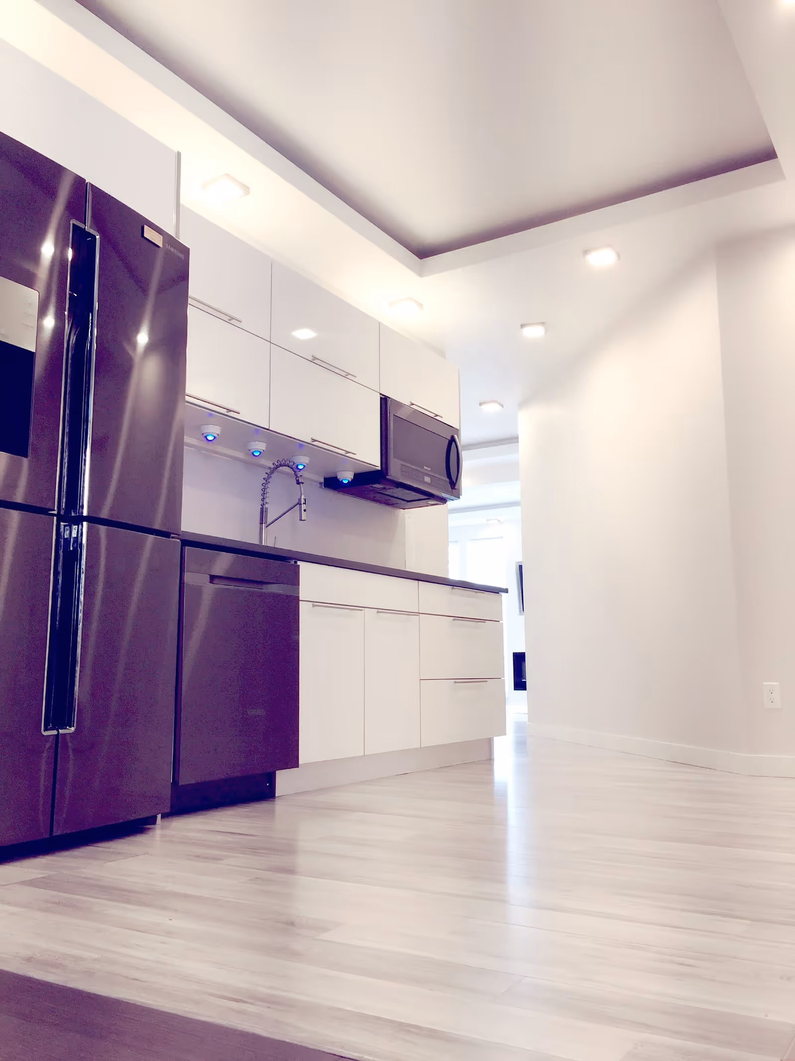 Modern kitchen with stainless steel refrigerator, dishwasher, and microwave. White cabinets with sleek handles, a black countertop, and a flexible kitchen faucet with blue LED lights underneath the upper cabinets. Light wood flooring and recessed ceiling lights.