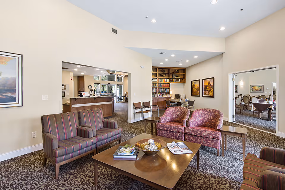 A bright assisted-living common lounge with upholstered sofas and armchairs around a coffee table, shelving and a dining area visible beyond.