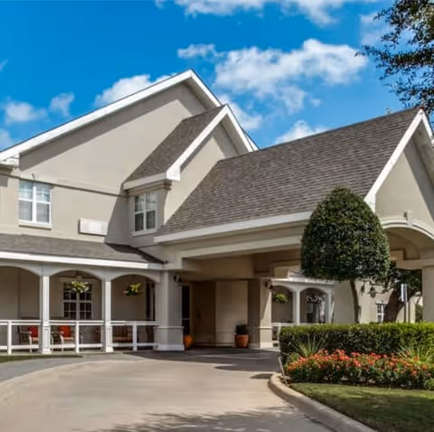 Exterior view of Parsons House Cypress senior living facility showing the entrance with a covered driveway, beige building with white trim, manicured bushes, flowers, and a clear blue sky with some clouds.