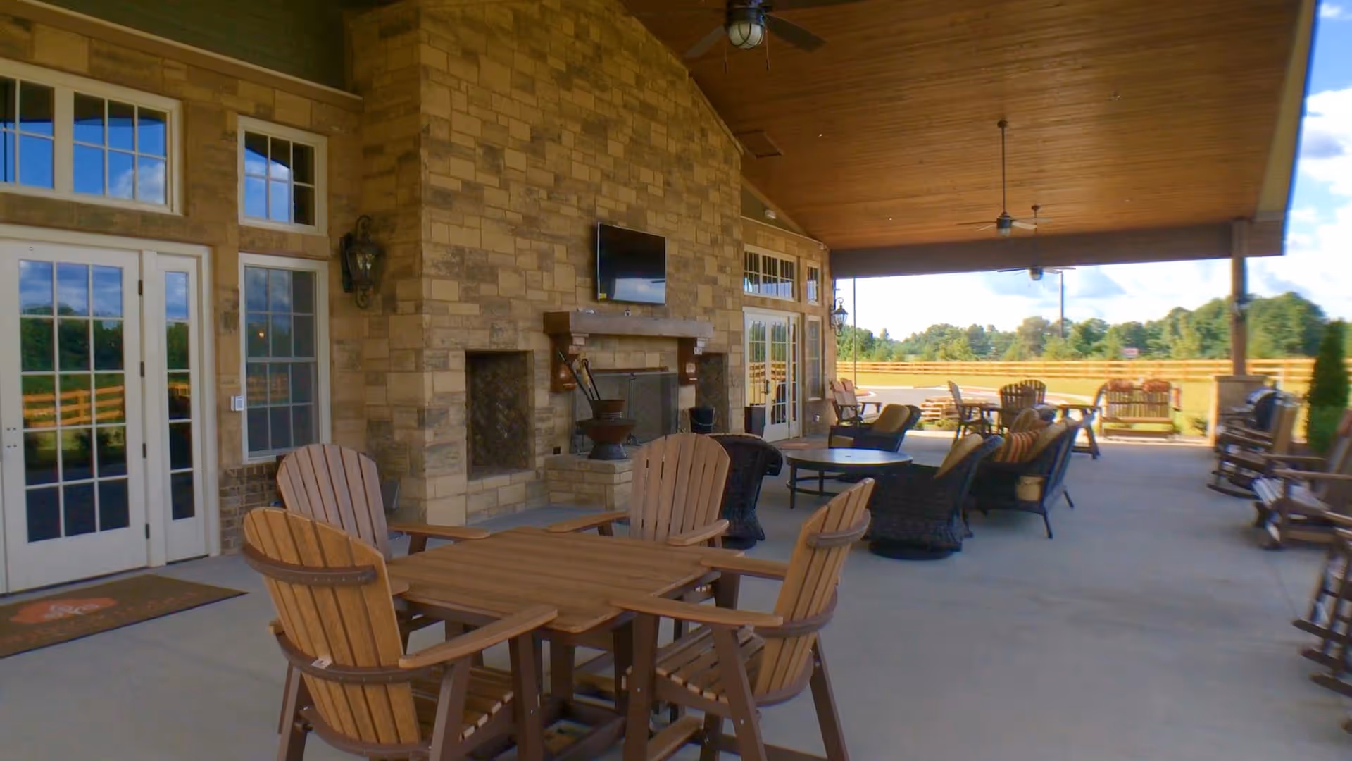 Covered outdoor patio area with wooden ceiling, stone fireplace, mounted TV, and multiple seating arrangements including wooden chairs around a table and cushioned lounge chairs. The patio overlooks a fenced grassy area with trees in the background under a partly cloudy sky.