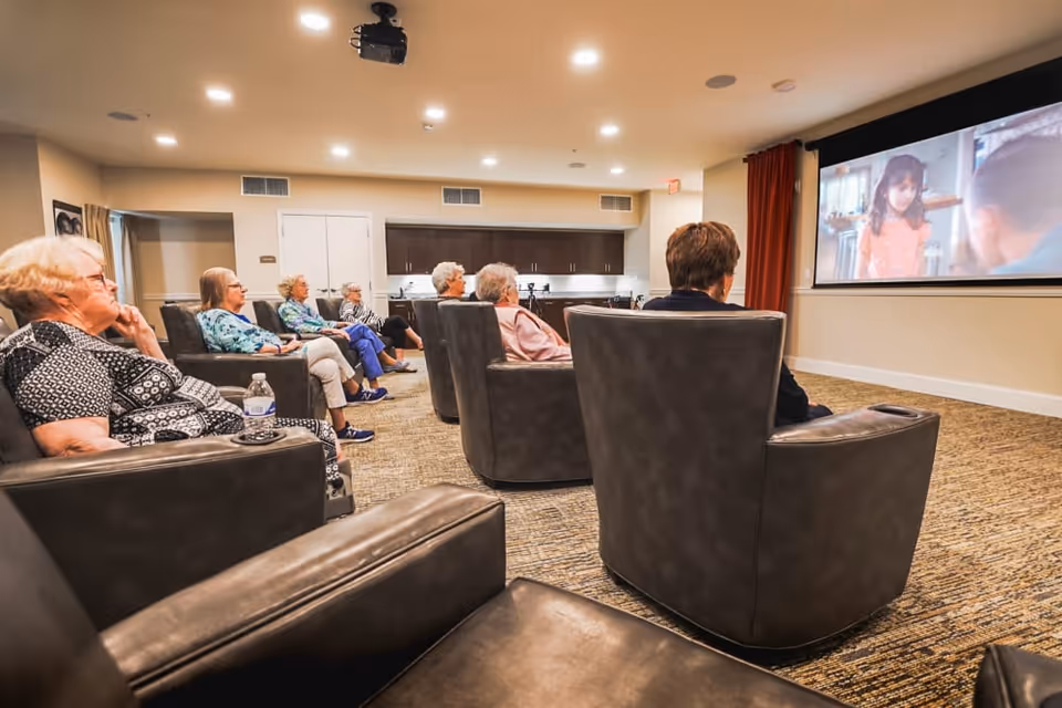 A group of elderly women seated in comfortable leather chairs watching a movie projected on a large screen in a cozy, well-lit common room with carpeted floor and a kitchenette in the background.