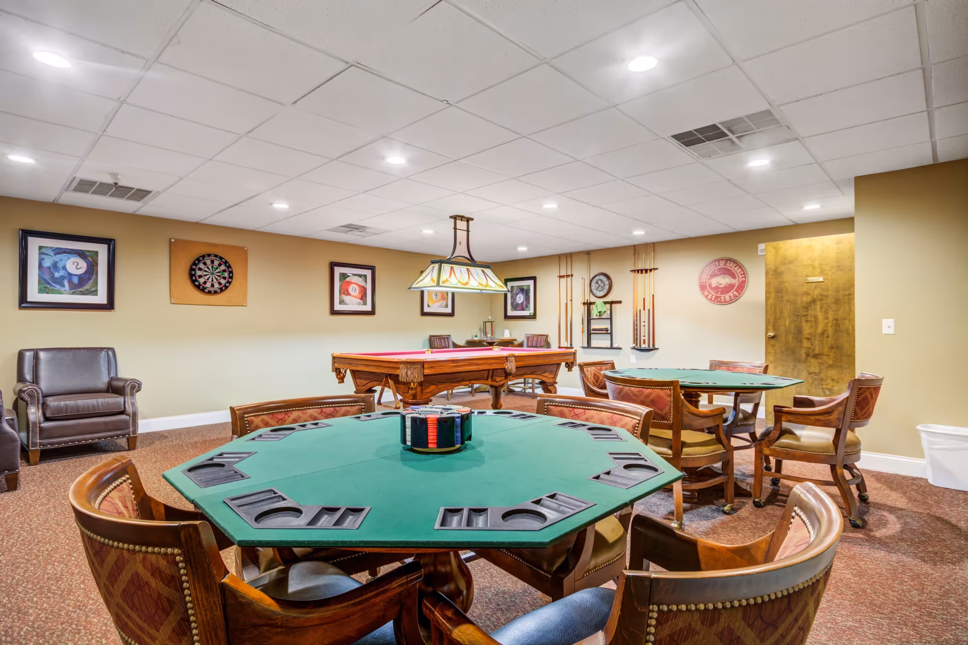 A recreational room with two green octagonal card tables surrounded by wooden chairs with patterned upholstery. In the background, there is a pool table with a red felt surface, a dartboard mounted on the wall, and several framed pictures of billiard balls. The walls are beige, and the ceiling has recessed lighting. There are also pool cues mounted on the wall and a wooden door on the right side.