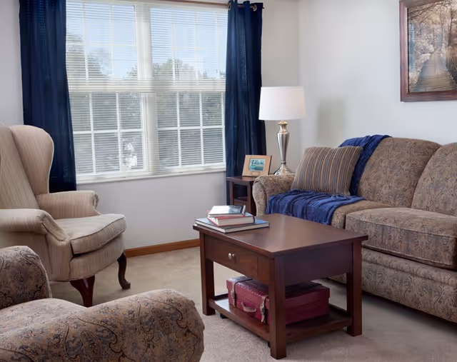 A cozy living room with a patterned sofa and armchair, a wooden coffee table with books on top, a side table with a lamp and framed photo, and a large window with blue curtains letting in natural light.