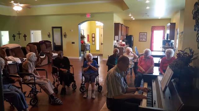 A group of elderly people seated in a common area, some in wheelchairs, listening to a man playing a piano. The room has yellow walls, brown recliners, and decorative crosses on one wall.