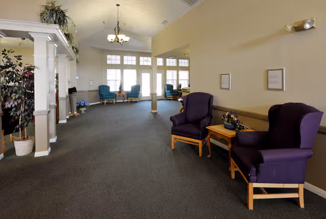 A spacious, well-lit common area with carpeted floor, beige walls, and large windows letting in natural light. The room features several armchairs in purple and teal colors arranged along the walls, small wooden tables with floral decorations, and potted plants near white columns on the left side.