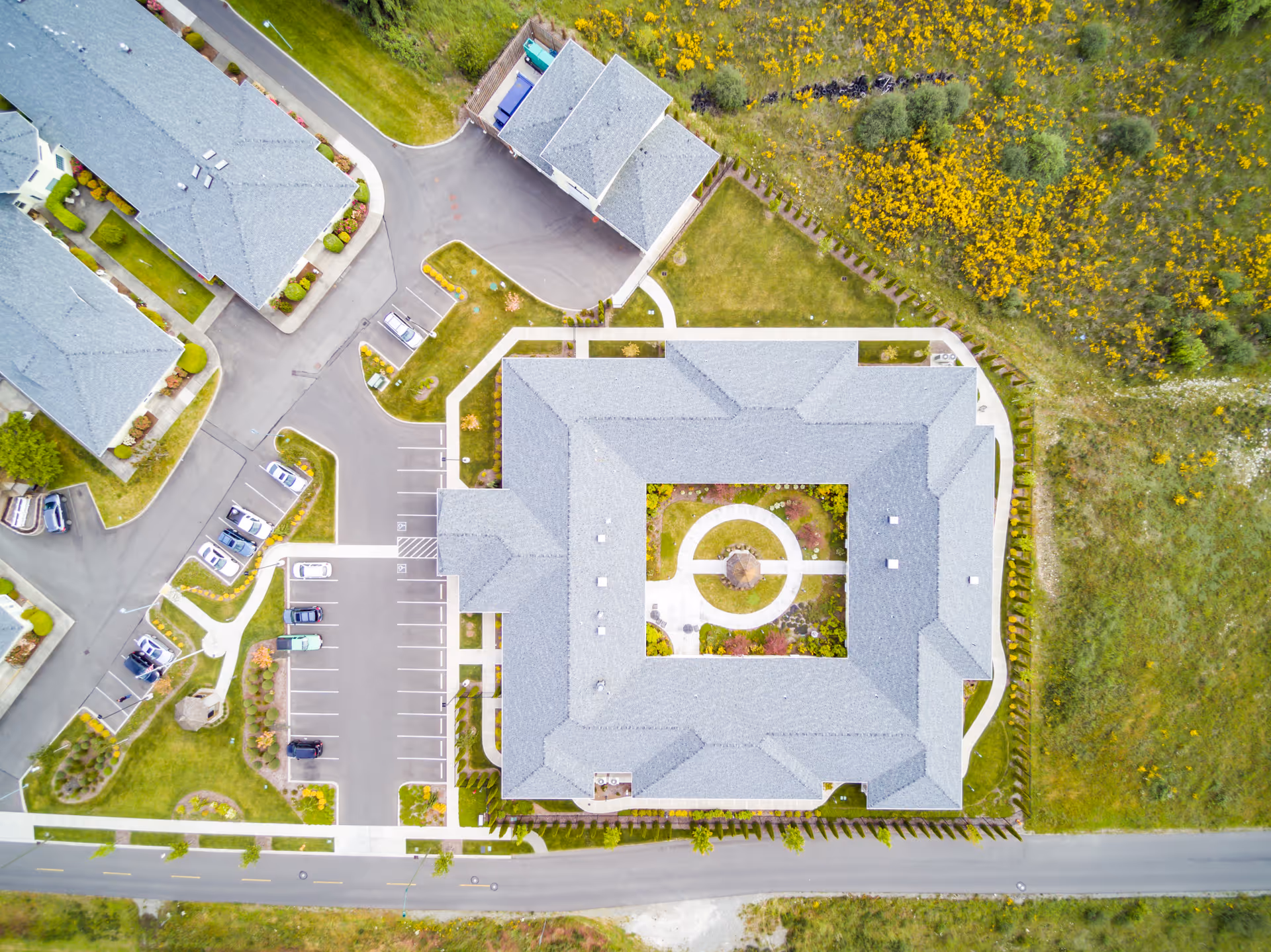 Aerial view of Easthaven Villa Assisted Living & Memory Care showing the building with a central courtyard, surrounding parking lots, adjacent smaller buildings, and landscaped areas with greenery and yellow flowers.