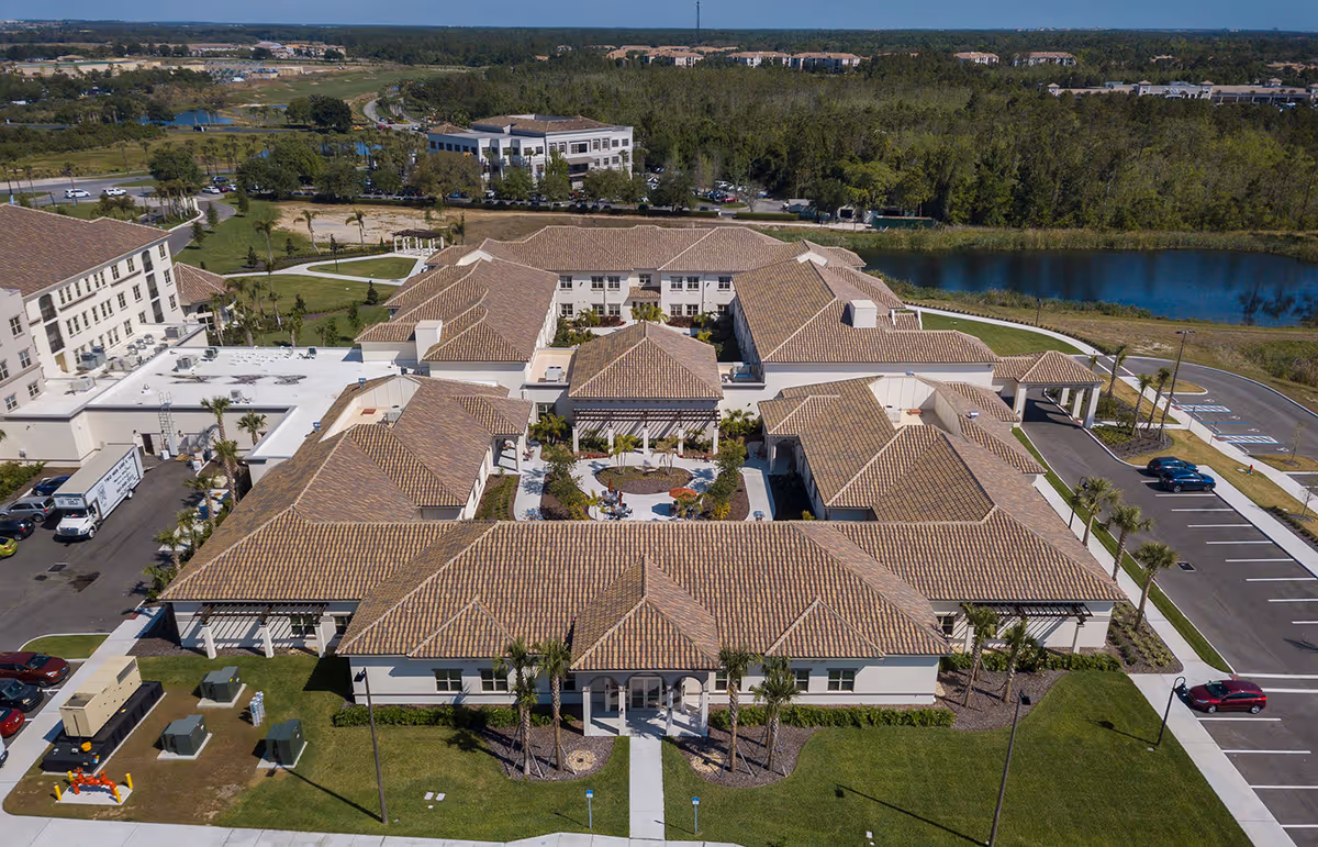 Aerial view of a Mediterranean-style senior living complex with terracotta tiled roofs surrounding a central courtyard, parking areas, and a nearby pond.