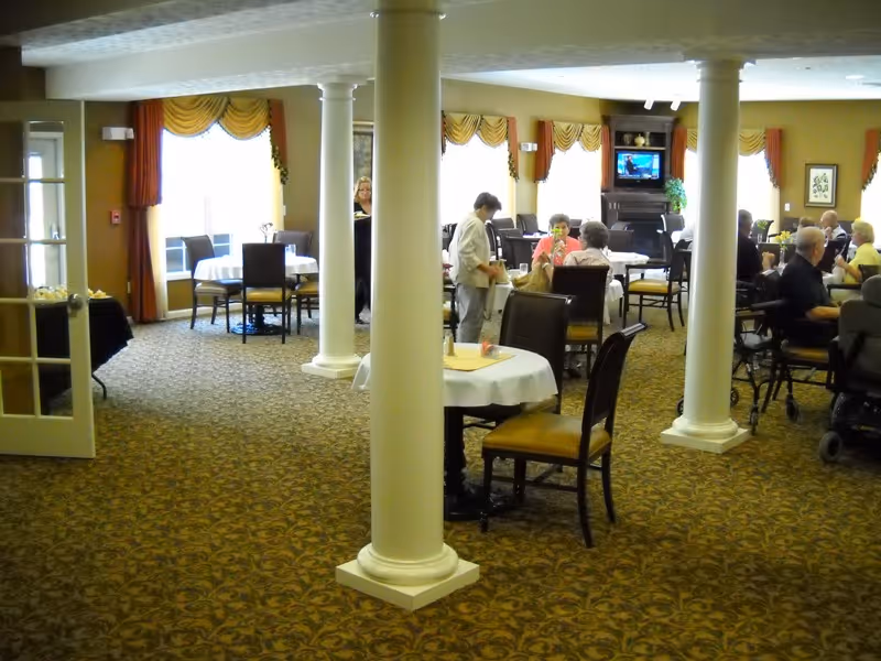 Assisted living dining room with round tables covered in white cloths, decorative columns, and residents seated and socializing.