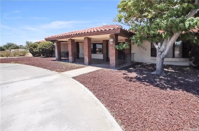 Exterior view of a single-story building with a covered porch supported by brick columns, a tiled roof, and a tree to the right. The ground is covered with reddish-brown mulch and there is a curved concrete driveway in front.