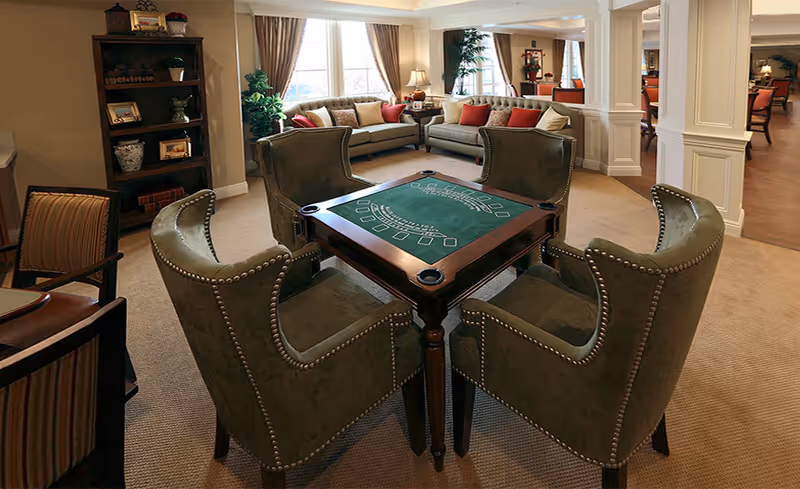 A cozy common area with a green card table surrounded by four upholstered chairs with nailhead trim. In the background, there are two sofas with cushions, a tall bookshelf with decorative items, a potted plant, and large windows with curtains letting in natural light. The room has beige carpeting and white columns separating different sections.