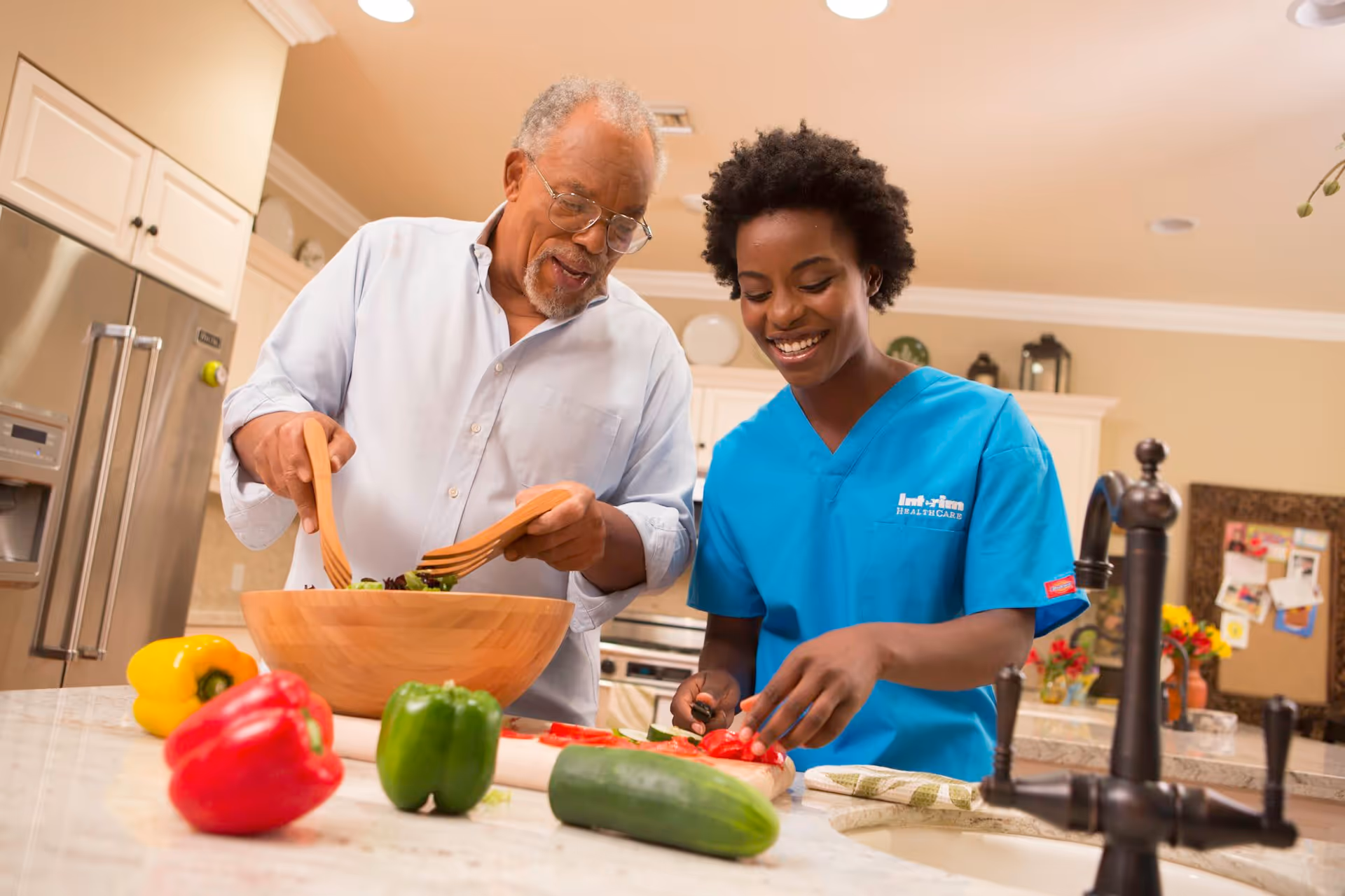An elderly man and a healthcare worker in blue scrubs are preparing a salad together in a kitchen. The man is tossing salad in a wooden bowl while the healthcare worker is chopping red bell peppers on a cutting board. Various vegetables including yellow, red, and green bell peppers and a cucumber are on the kitchen counter.