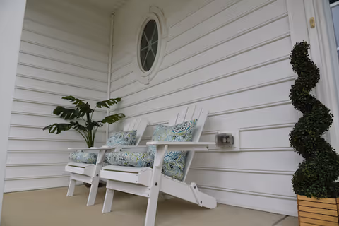 White exterior wall with circular window, two white chairs, and a topiary on the porch.