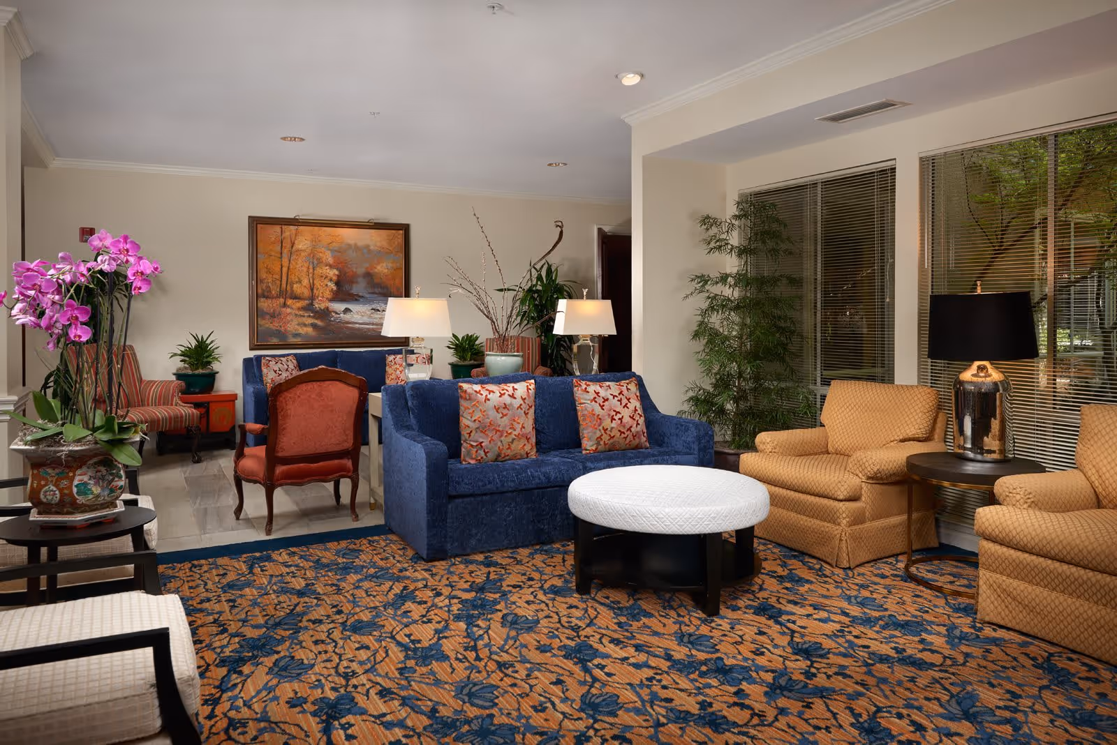 A cozy common lounge with a blue sofa, upholstered armchairs, a round white ottoman, patterned rug, plants, lamps, and framed artwork.