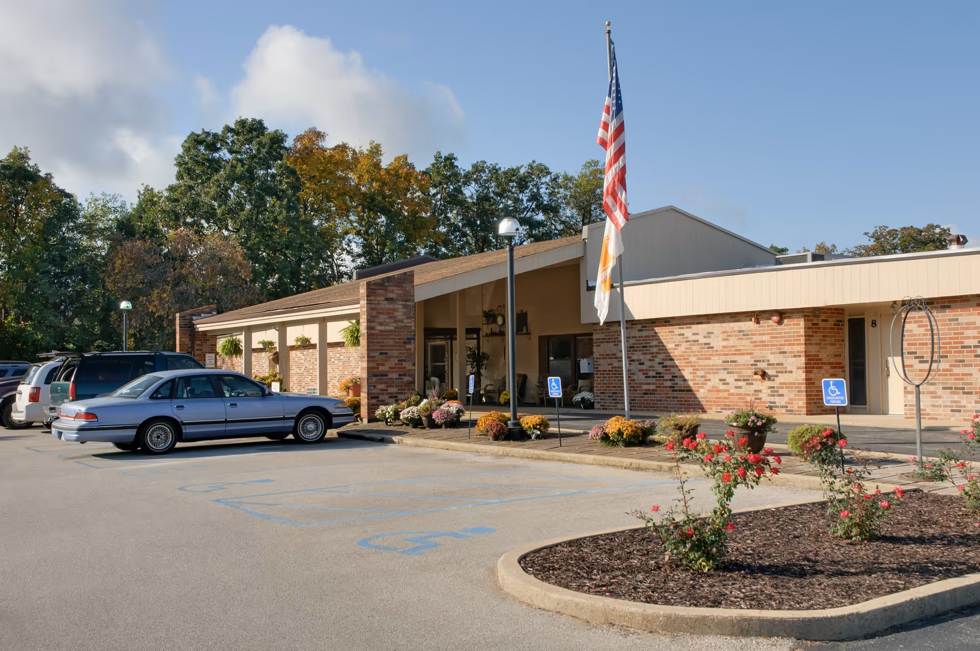 Entrance and parking area of a single-story brick senior care facility with an American flag, cars, and landscaped flower beds.