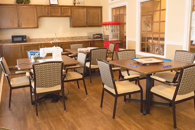 A room with multiple tables and chairs arranged for group activities. One table has a sewing machine and fabric, while another table has a board game set up. The room has wooden cabinets, a microwave, a popcorn machine, and a countertop along one wall. The floor is wooden, and there are windows with wooden frames on one side.