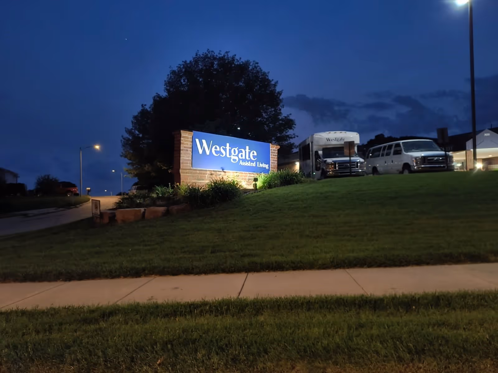 Illuminated sign for Westgate Assisted Living on a grassy hill at dusk with a sidewalk in the foreground and several vehicles, including a shuttle bus, parked near the building in the background.