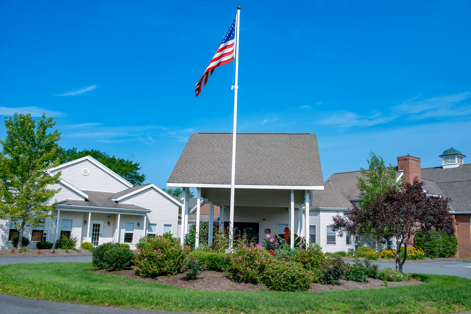 Front entrance of a senior living facility featuring a tall American flagpole, covered porte-cochere, and landscaped circular driveway.