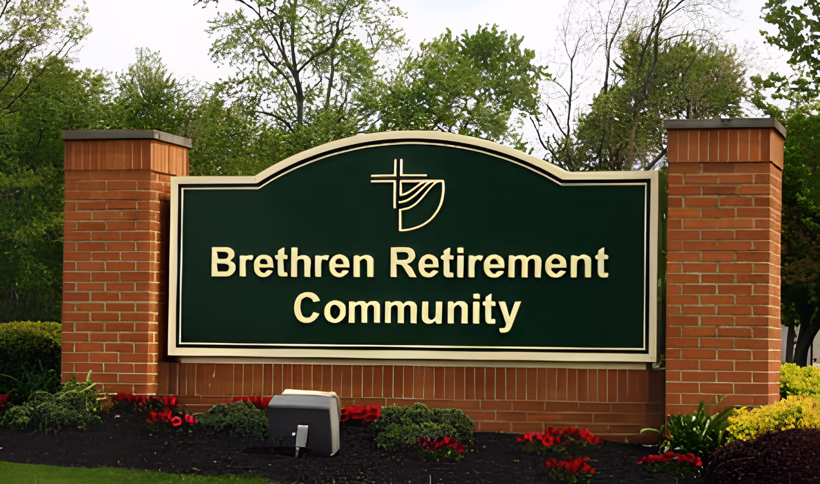 A large green sign with gold lettering reading 'Brethren Retirement Community' mounted between two brick pillars, surrounded by landscaped bushes and flowers with trees in the background.