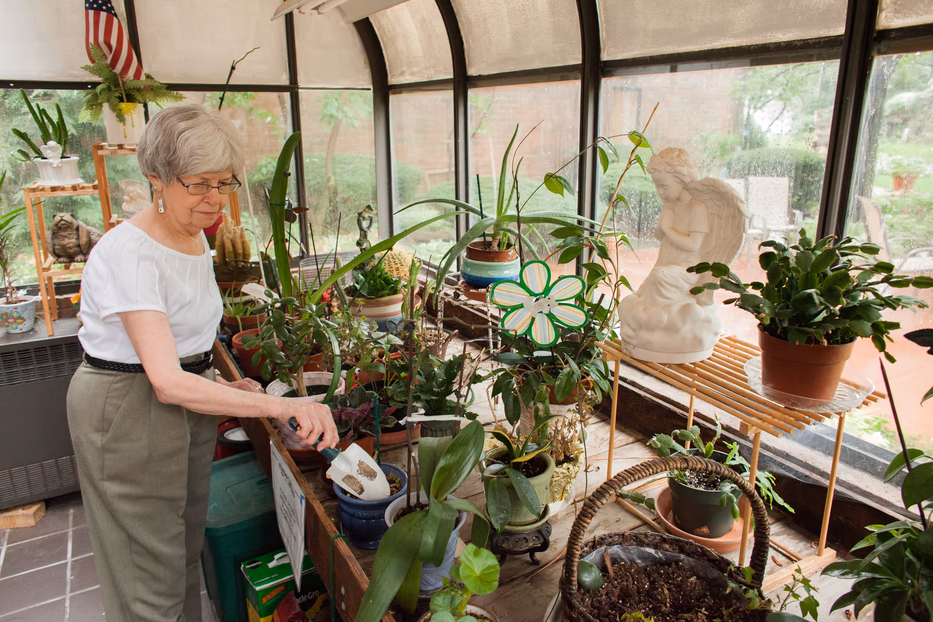 An elderly woman with short gray hair and glasses is tending to various potted plants inside a sunroom or greenhouse area. The space is filled with green plants, a white angel statue, and gardening tools. Large windows provide a view of the outdoor garden area.