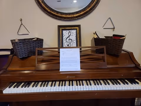 A wooden piano with a sheet of music on the stand, two triangle percussion instruments hanging on the wall above it, two dark woven baskets on top of the piano, and a framed musical note artwork between the baskets. A round mirror is partially visible above the framed artwork.
