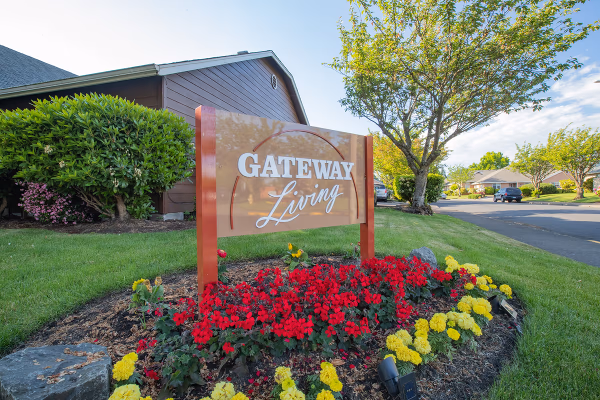 A landscaped outdoor area featuring a sign that reads 'Gateway Living' surrounded by red and yellow flowers, with a tree and residential buildings in the background under a clear sky.