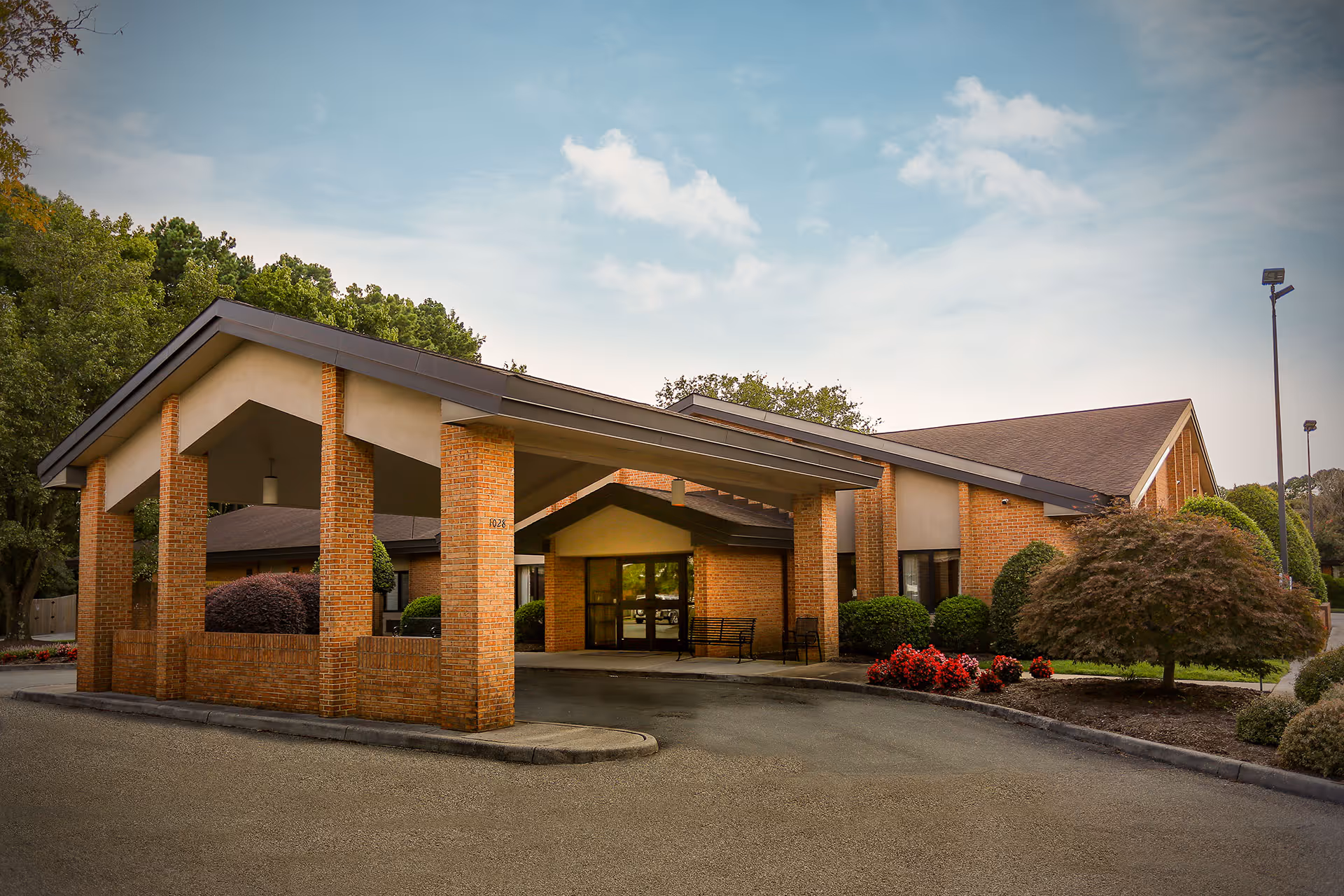 Exterior view of Northampton Nursing & Rehabilitation Center showing a brick building with a covered entrance supported by brick pillars, surrounded by landscaped bushes and trees under a partly cloudy sky.