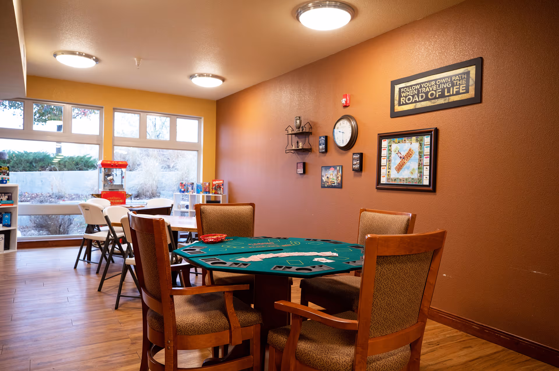 A cozy game room with a green octagonal card table surrounded by four wooden chairs with patterned cushions. The walls are painted in warm tones, with framed pictures and a clock hanging on the wall. In the background, there are folding chairs and tables near large windows letting in natural light, along with shelves holding board games and a popcorn machine.