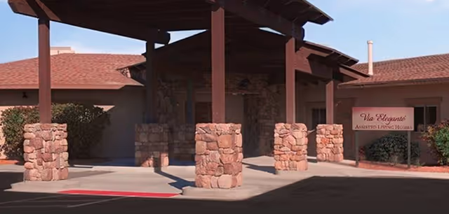 Covered main entrance of an assisted living facility with wooden beams, stone pillars, and a sign reading 'Via Elegante Assisted Living Homes'.