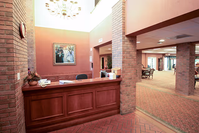 Reception area with a wooden front desk, a chair behind the desk, a telephone, and some office supplies. The walls are painted in a warm tone with brick columns and a framed painting hanging on the wall. The space is well-lit with a chandelier and opens into a larger room with carpeted flooring and several tables and chairs.
