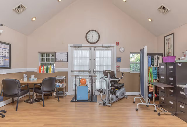 A room with light beige walls and wooden flooring featuring a table with chairs on the left, exercise equipment including a stationary bike and a balance trainer in the center, and a filing cabinet with organized items on the right. There are two windows and double doors with sheer curtains in the background, a clock above the doors, and various posters and charts on the walls.