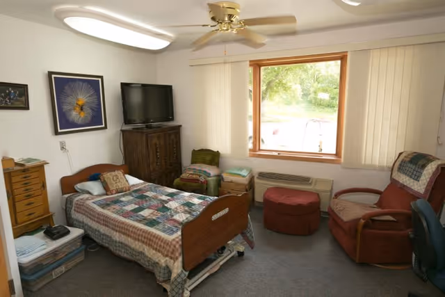 A single bedroom in a senior living facility with a bed covered in a patchwork quilt, a wooden cabinet with a TV on top, a red armchair with a quilt draped over it, a small round ottoman, a window with vertical blinds, and a ceiling fan with a light fixture.