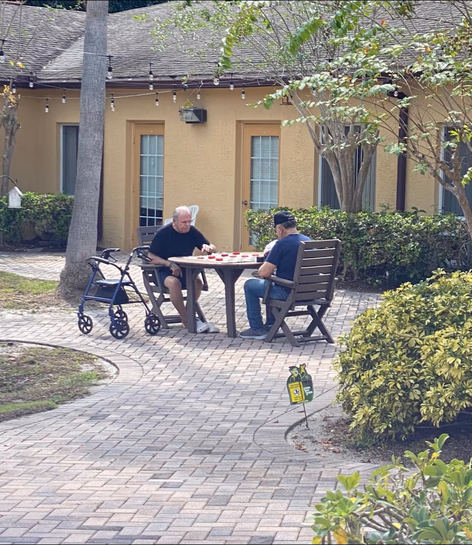 Two elderly men sitting at a wooden table outside on a paved patio, playing a board game. One man has a walker next to him. The background shows a beige building with doors and windows, surrounded by bushes and trees.