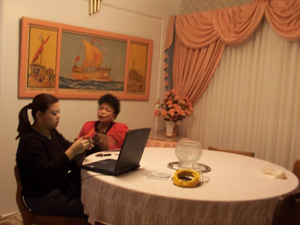 Two people sit at a round dining table with a laptop, a glass centerpiece, and a floral arrangement beneath framed artwork and draped curtains.