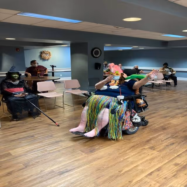 A group of elderly people sitting in a common room with blue walls and wooden floors. One person in a wheelchair is dressed in a colorful hula skirt and a flamingo hat, holding a green inflatable alligator. Another person is dressed as a witch with green face paint and a black hat. Other individuals are seated on chairs along the walls, some wearing masks.