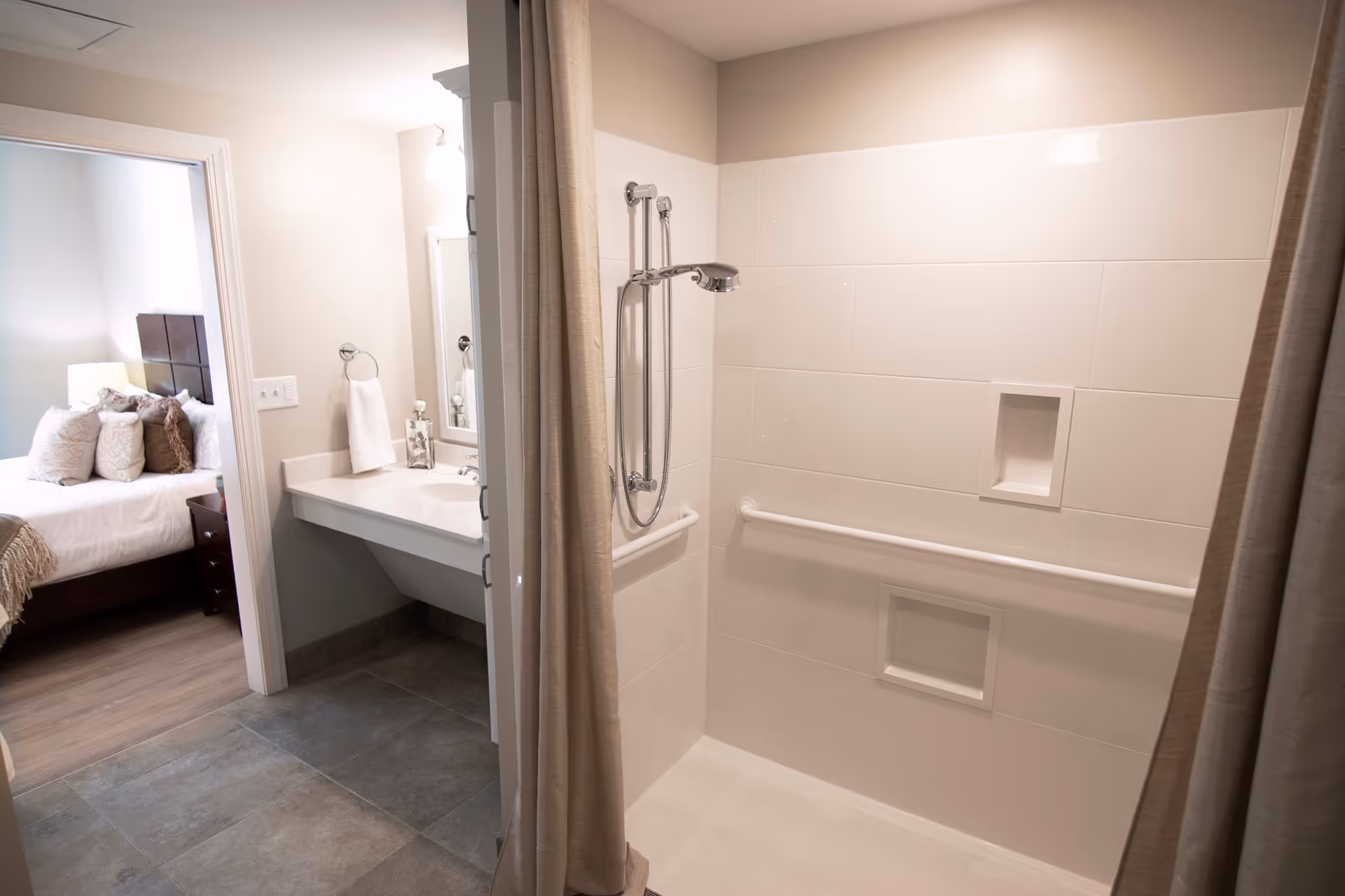 A bathroom with a walk-in shower featuring a handheld showerhead and grab bars. The shower has built-in shelves and a beige curtain. Adjacent to the shower is a sink with a mirror and a towel ring holding a white towel. In the background, a bedroom with a bed, pillows, and a nightstand with a lamp is visible.