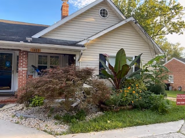 Exterior view of a single-story house with white siding and a brick chimney. The front yard features a variety of plants and shrubs, including a large leafy plant and a small tree. A sidewalk runs along the front of the house, and another brick building is visible in the background.