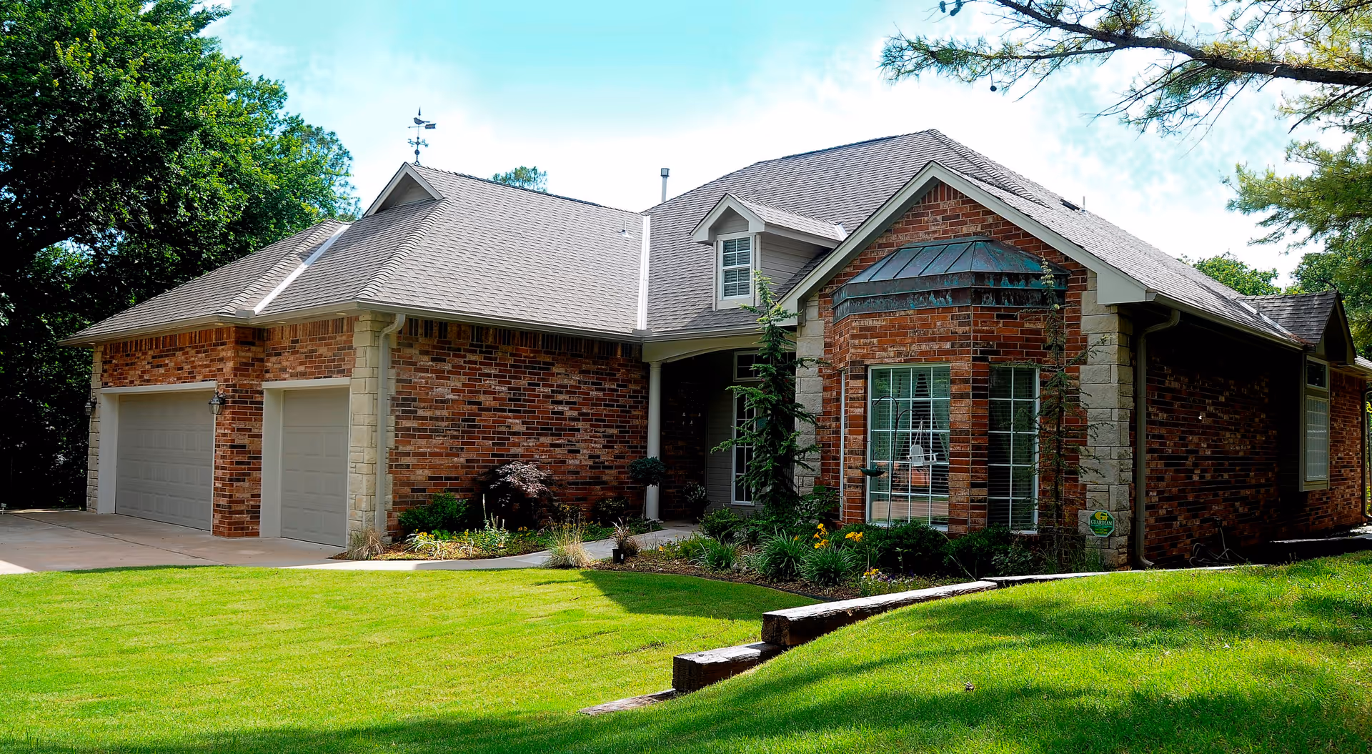 A single-story brick house with a gray shingled roof, large windows, and a well-maintained green lawn with some shrubs and trees around the property under a blue sky.