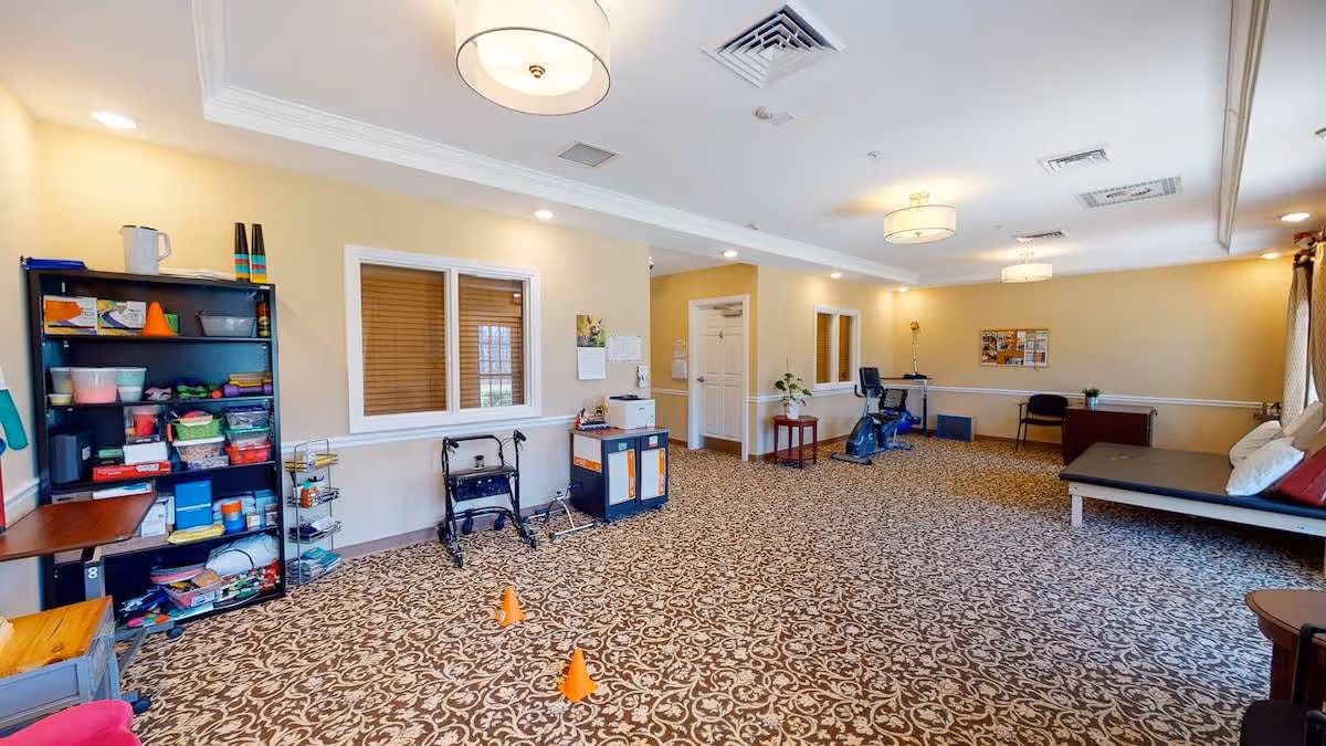 A spacious room with patterned carpet and beige walls, featuring a black shelving unit with various supplies, two orange cones on the floor, a walker, a stationary exercise bike, a padded bench with pillows, a desk with a chair, and ceiling lights.