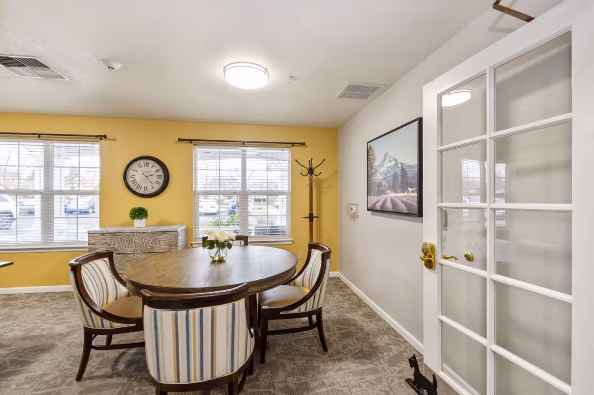 A bright dining room with a round wooden table surrounded by four striped upholstered chairs. The walls are painted yellow and white, with two large windows letting in natural light. A wall clock and a small plant sit on a wooden cabinet beneath the windows. A coat rack stands in the corner, and a framed picture of a mountain landscape hangs on the white wall. A glass-paneled door with a brass handle is partially open, revealing more of the room.