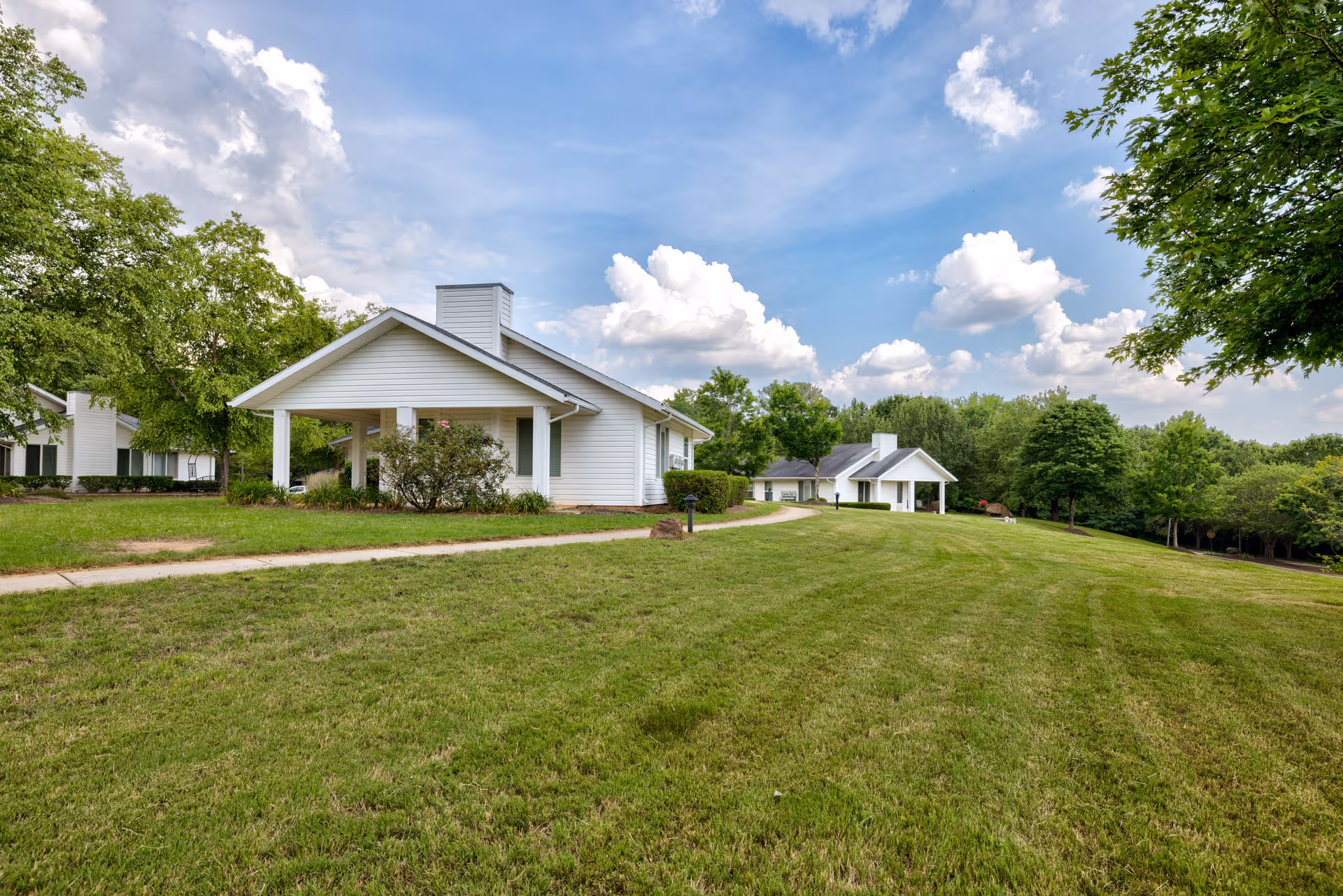 A scenic view of a senior living facility with white single-story buildings surrounded by green lawns and trees under a partly cloudy blue sky.