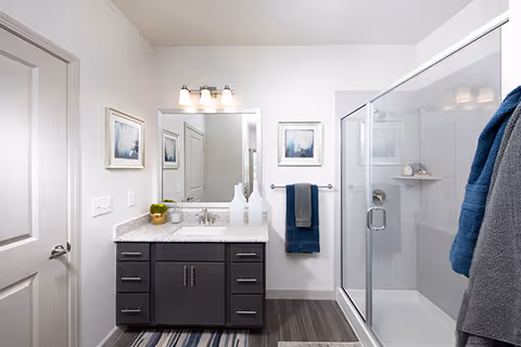 A modern bathroom featuring a glass-enclosed shower on the right, a dark wood vanity with a granite countertop and a sink in the center, a large mirror above the vanity with three light fixtures, two framed pictures on the wall, and a towel rack holding blue and gray towels.
