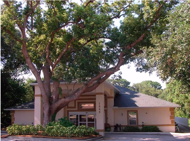 Exterior view of a single-story building with beige walls and a dark roof, partially shaded by a large tree with green leaves in front. The building has a glass entrance door and windows, surrounded by landscaping with bushes and plants.