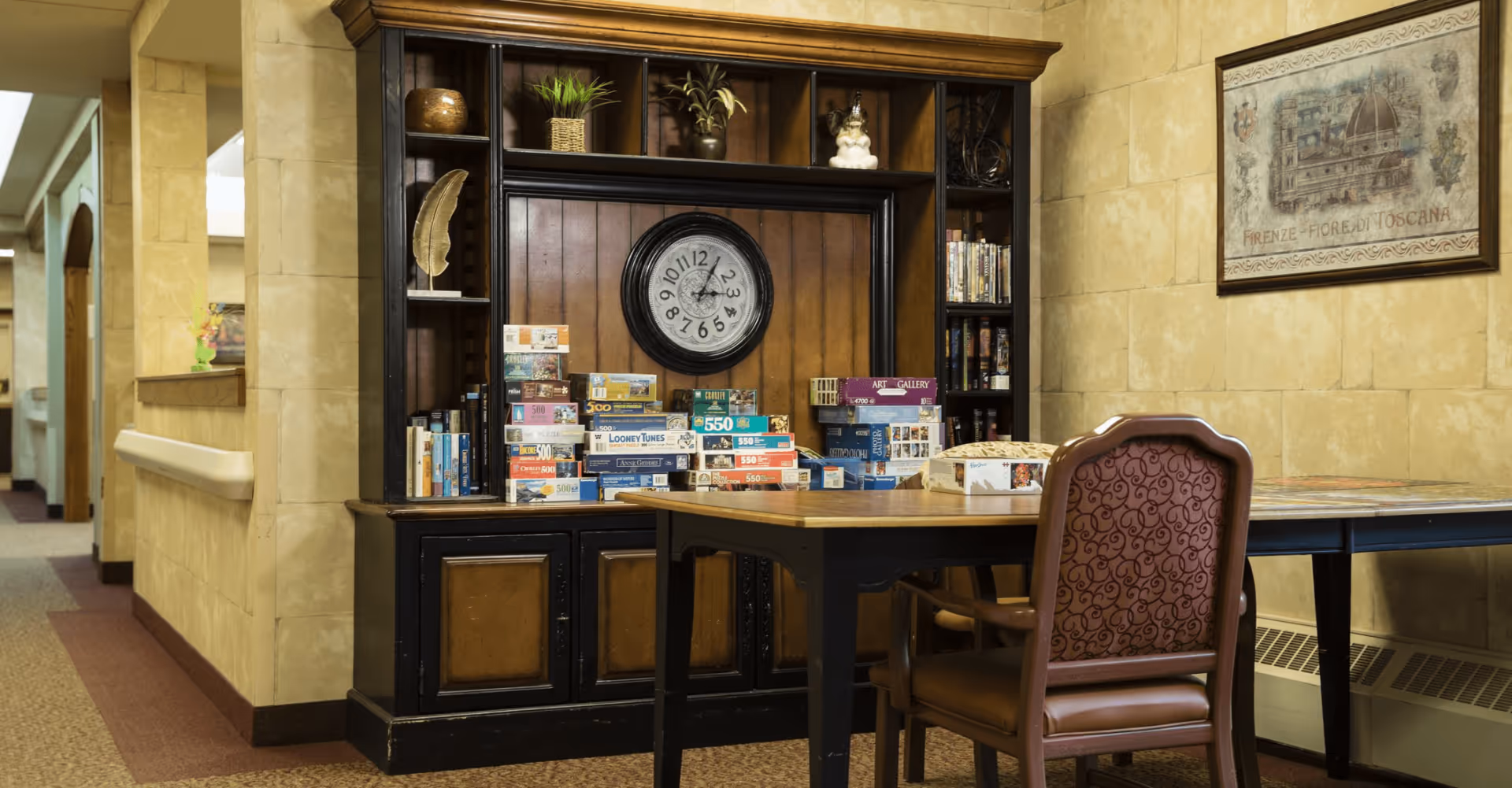 A cozy communal activity area with a wooden shelving unit holding a clock and stacked puzzle boxes, and a table with an upholstered chair in front.