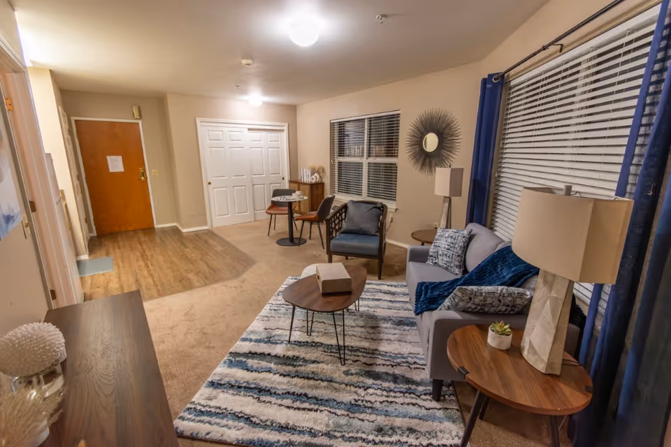 A cozy living room area with a gray sofa adorned with patterned pillows and a blue throw blanket. There are two wooden side tables with lamps and a small plant. A patterned rug covers the carpeted floor, and a wooden coffee table sits in the center. A single armchair is positioned near a small round table with two chairs by a window with blinds. The room has beige walls, a wooden floor near the entrance door, and a decorative round mirror on the wall.